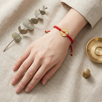 A hand wearing a red braided string bracelet with a gold coin charm displaying Chinese characters rests on a neutral linen surface next to a dried eucalyptus branch, a brass thimble, and a small brass dish holding a gold ring