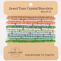 Cherry Tree Collection Jewel Tone Crystal Bracelets a set of twelve faceted crystal bead bracelets with gold accents displaying an ombre color gradient from peach and pink to cool blues and greens on a kraft paper display card
