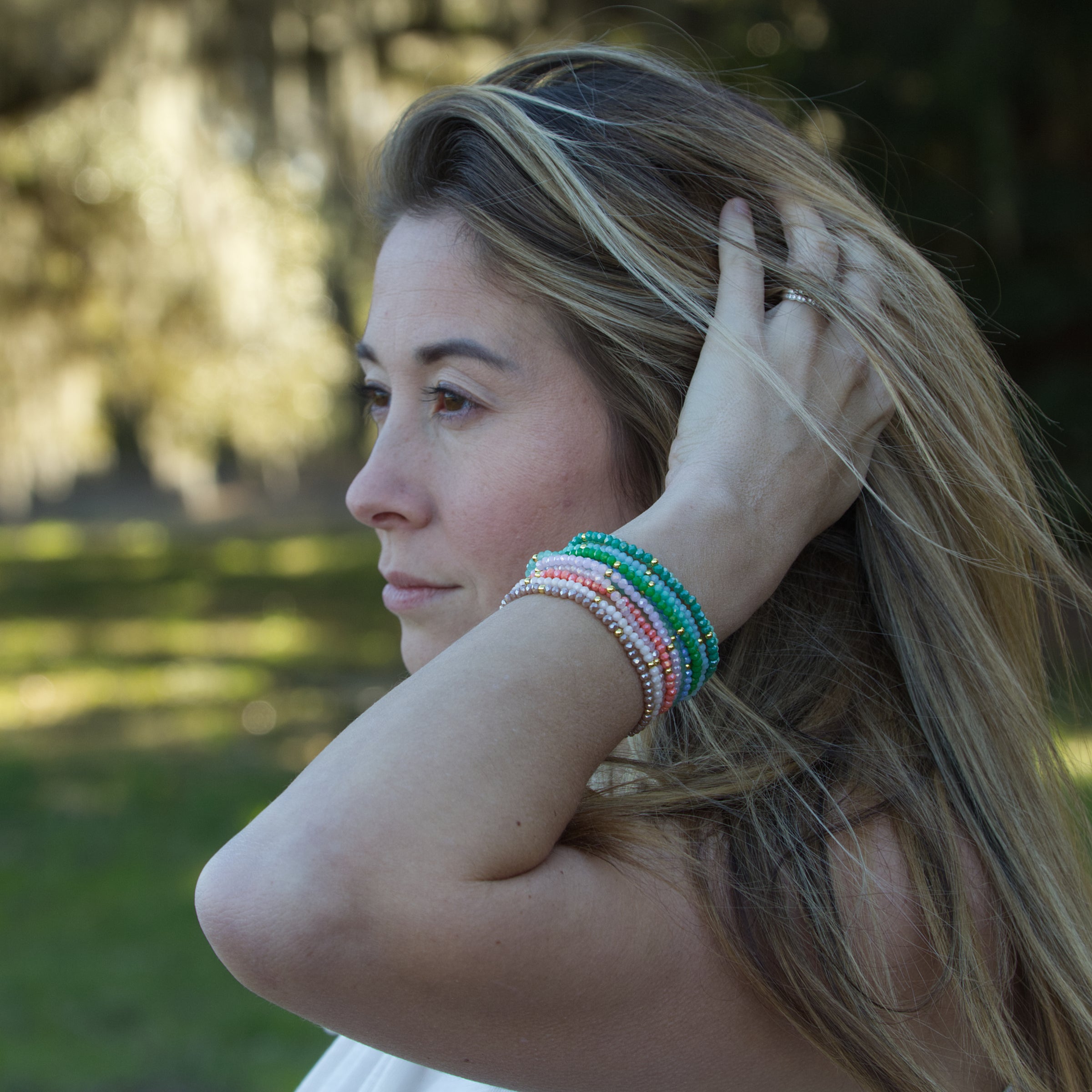 A woman with blonde highlighted hair running her hand through her locks, adorned with a stack of pink, green, and nude beaded bracelets, looking thoughtfully into a blurry natural background