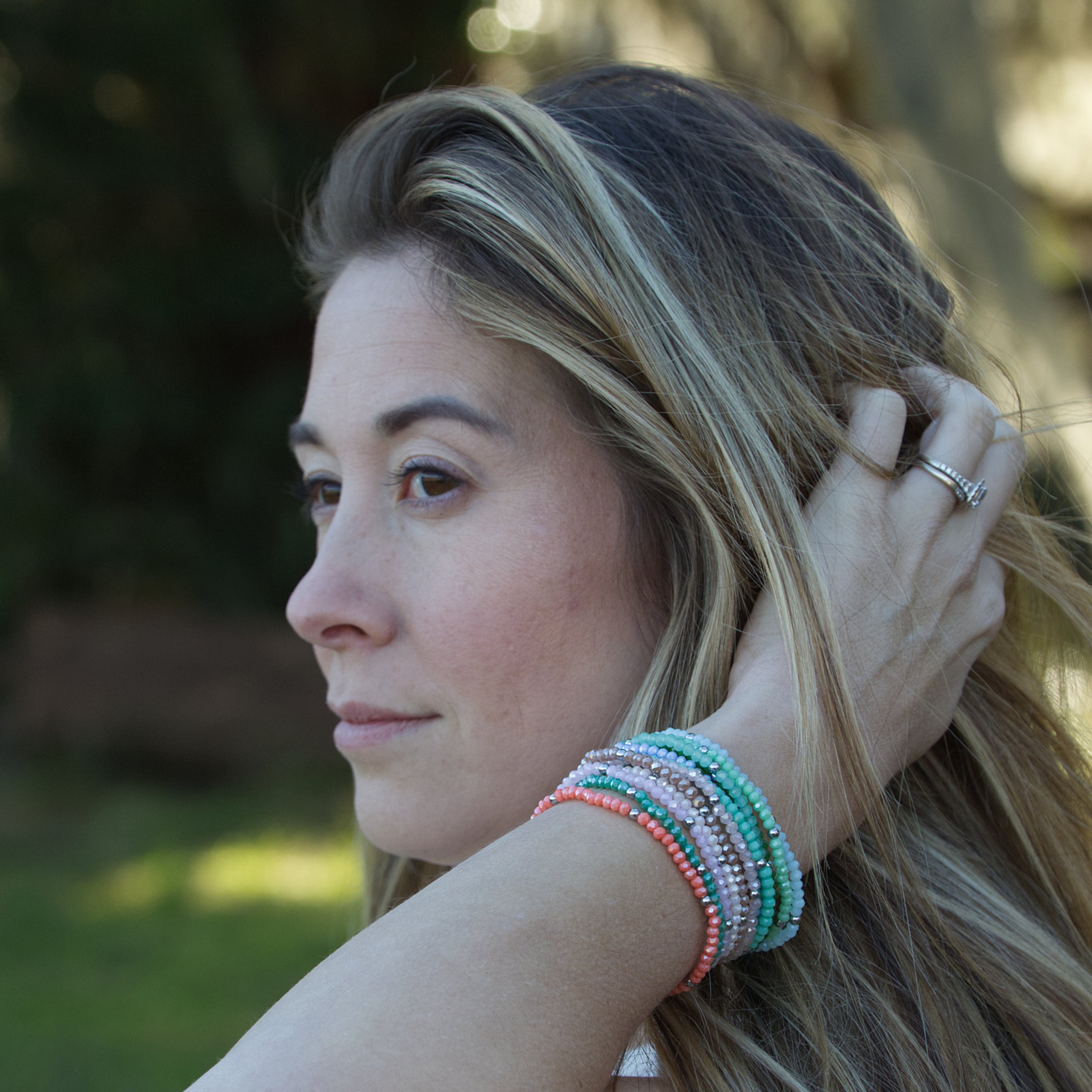 A woman in profile with highlighted blonde hair and a stack of colorful coral pink mint and silver beaded bracelets, her left hand with a diamond ring gently touching her hair outdoors