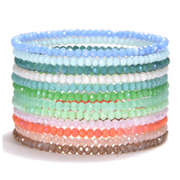 A vibrant stack of multiple beaded bracelets crafted from small faceted glass beads displaying a color gradient from cool blues and greens to warm pinks coral and brown on a white background