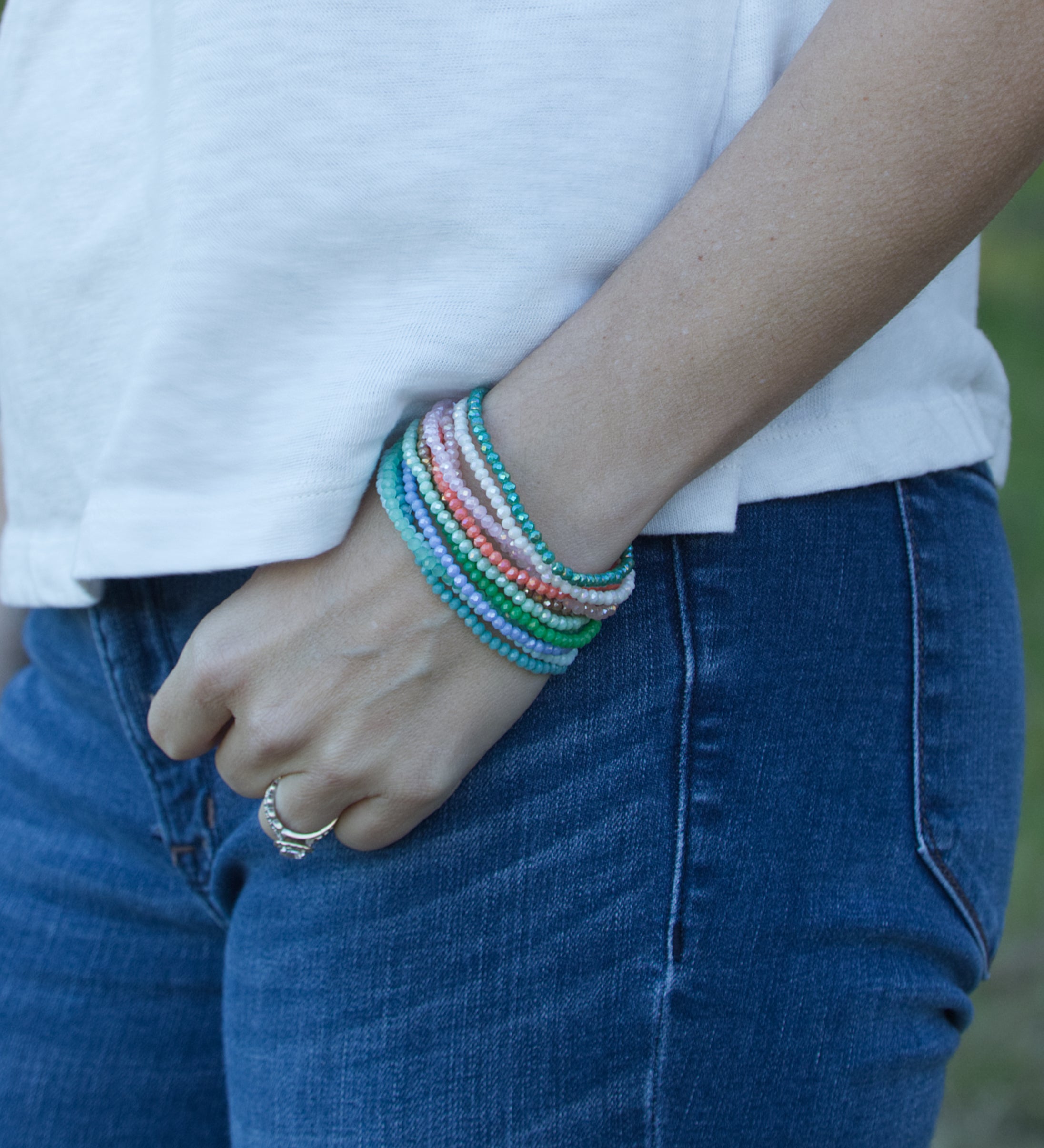 Close-up photograph of a person wearing a white t-shirt and blue denim jeans, featuring a stack of vibrant beaded bracelets in blues, greens, pinks, and purples on their wrist, with a gold diamond ring on their finger against a soft green outdoor background
