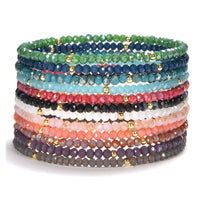 A vibrant stack of faceted crystal bead bracelets in a spectrum of colors including green blue red black white pink coral and purple accented with small gold spacer beads on a clean white background