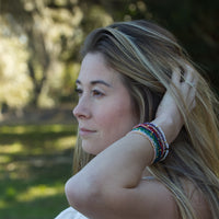Photograph of a woman in profile looking left with blonde-streaked hair and a stack of colorful beaded bracelets on her left wrist, wearing a diamond ring, against a soft blurred green outdoor background