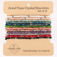 Cherry Tree Collection beaded bracelets: a set of twelve Jewel Tone Crystal Bracelets on a kraft paper display card, showcasing faceted crystal beads in a rainbow of hues like pastel pink, deep teal, and bright red, accented with small silver-colored spacer beads, and featuring the brand's "Handmade To Inspire" text