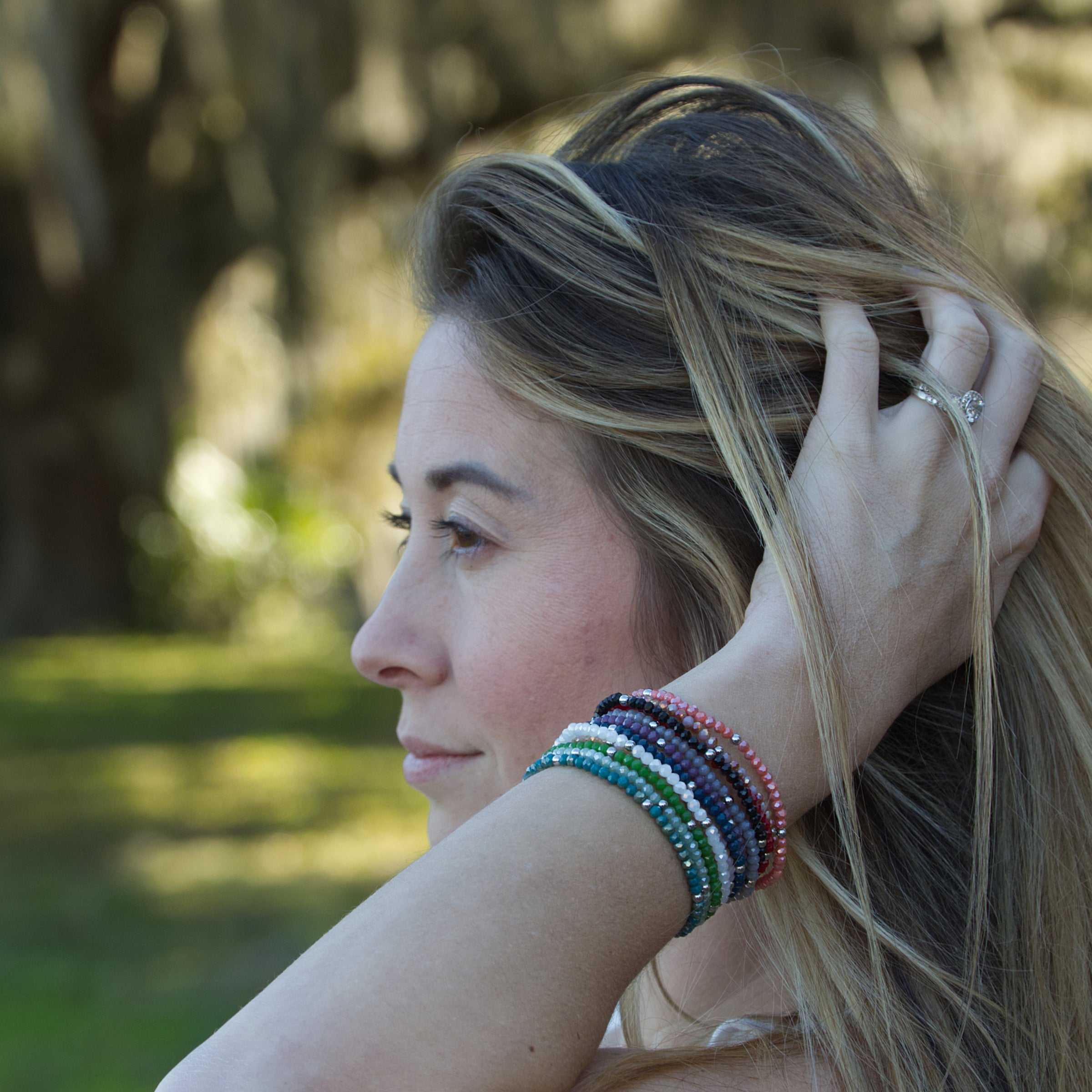 Photography of a woman in profile outdoors her hand running through her highlighted blonde hair showcasing a diamond engagement ring and a stack of colorful beaded bracelets on her wrist