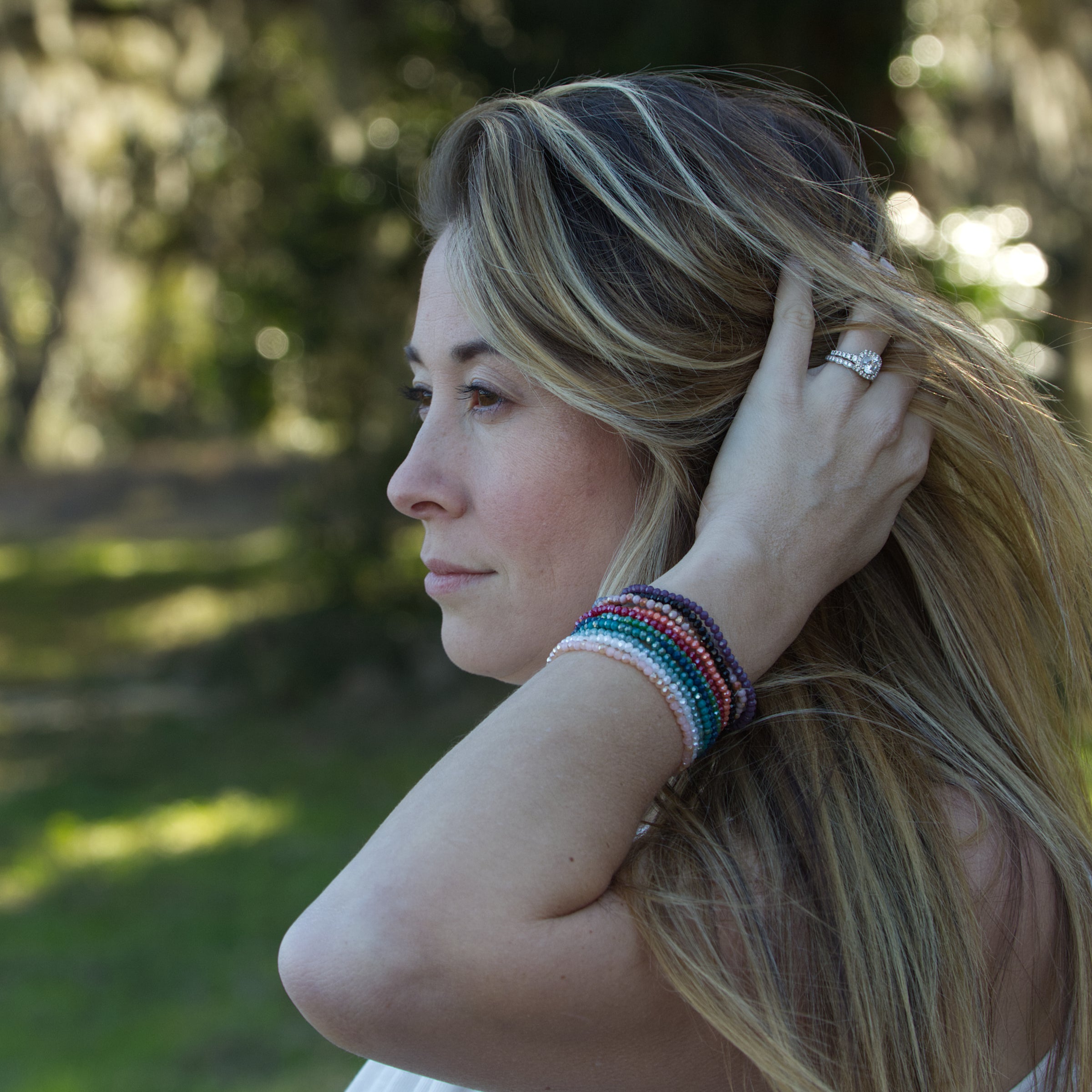 A close-up photograph of a woman with long blonde highlighted hair, wearing a prominent diamond engagement ring and a vibrant stack of multi-colored beaded bracelets, looking off-camera against a soft green bokeh background