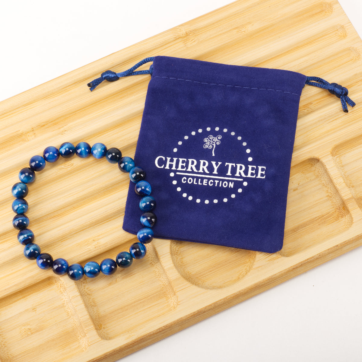 Product photography of a blue tiger eye beaded bracelet next to a navy blue velvet Cherry Tree Collection drawstring pouch on a light brown bamboo serving tray with shallow indentations