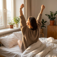 Photograph of a woman in a beige sweater stretching her arms upward in bed with sunlight streaming through a window illuminating potted green plants on the sill and a red beaded bracelet on her wrist