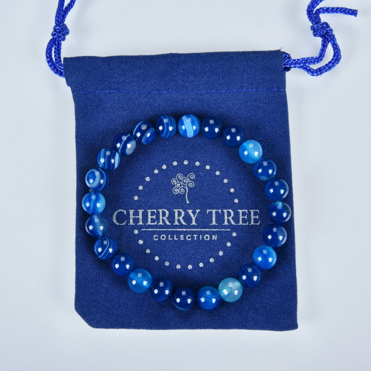 A vibrant blue agate beaded bracelet with both solid and banded beads laid on a navy blue drawstring pouch featuring a silver Cherry Tree Collection logo and dotted border